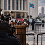 Caméras et micros devant un palais officiel à Kinshasa, drapeau congolais. Photo TIC-TAC HEBDO générée par intelligence artificielle — illustration, pas un reportage.
